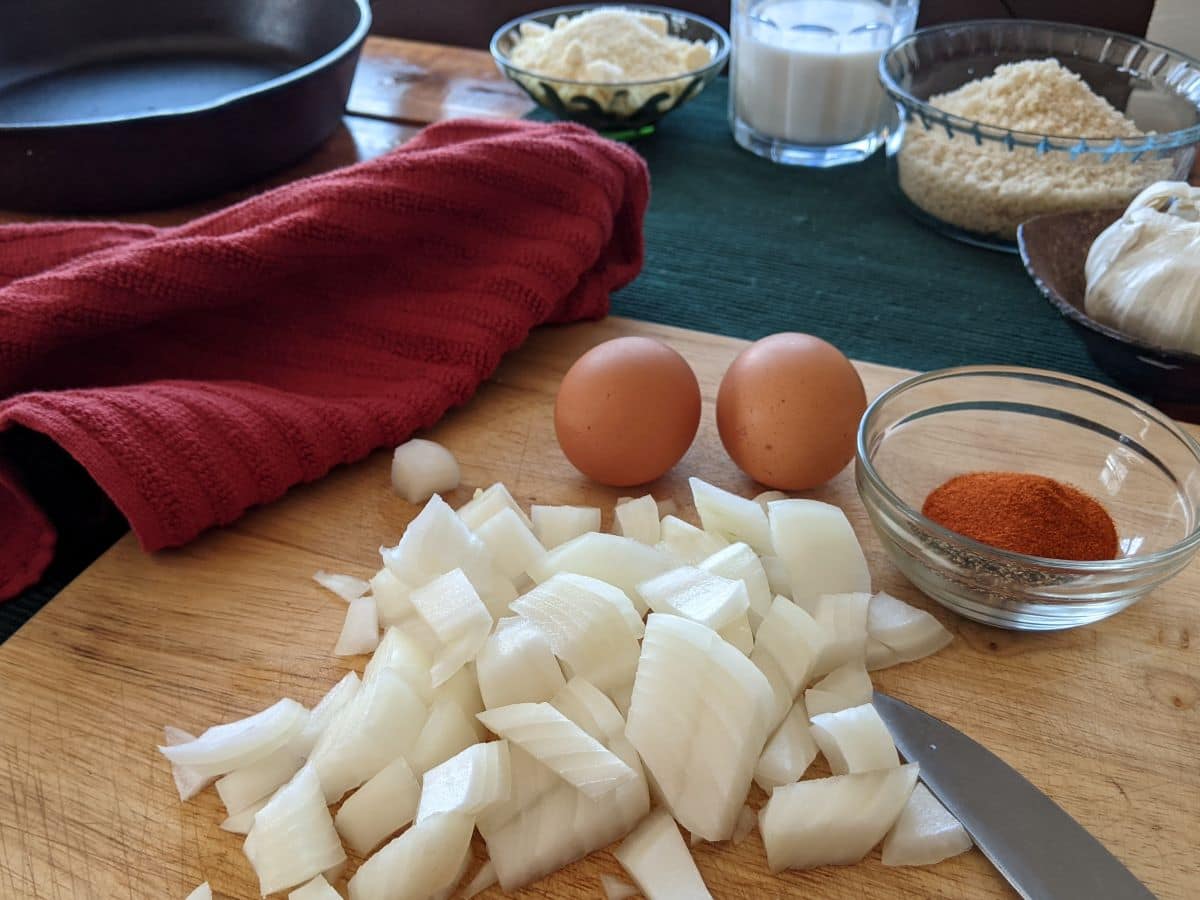 Chopped onions, two whole eggs, a bowl of paprika, and a knife sit on a wooden cutting board. In the background are a red cloth, skillet, bowls of breadcrumbs, grated cheese, milk, and a head of garlic.