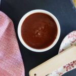 A bowl of dark red barbecue sauce sits on a black slate surface with a red and white cloth nearby.