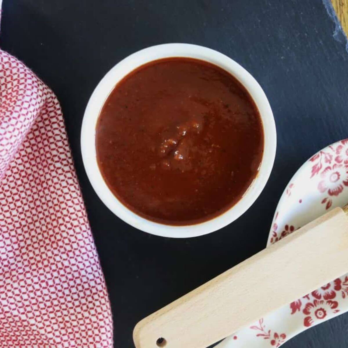 A bowl of dark red barbecue sauce sits on a black slate surface with a red and white cloth nearby.