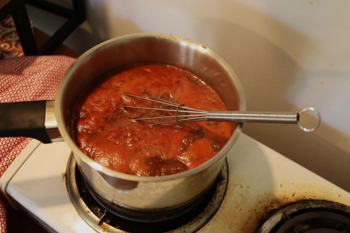 Red bbq sauce simmering in a small saucepan with a whisk in it on a stovetop.