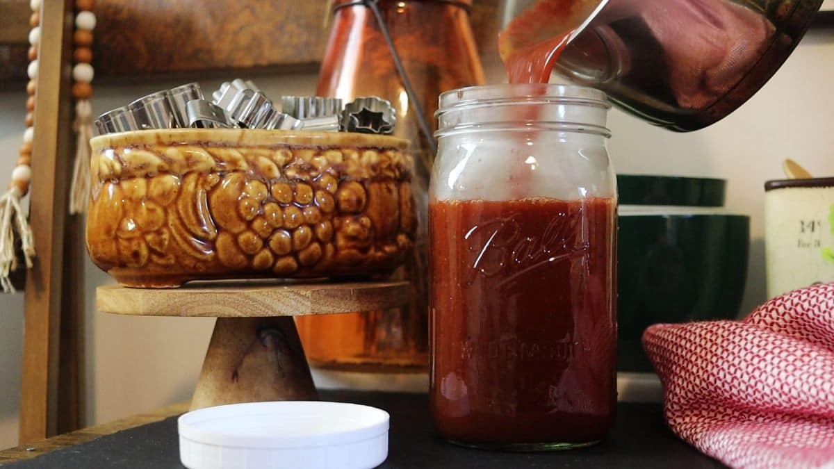 Pouring bbq sauce from a small saucepan into a quart size mason jar sitting on a countertop with a white lid and a red and white cloth next to it, there are kitchen utensils in the background.