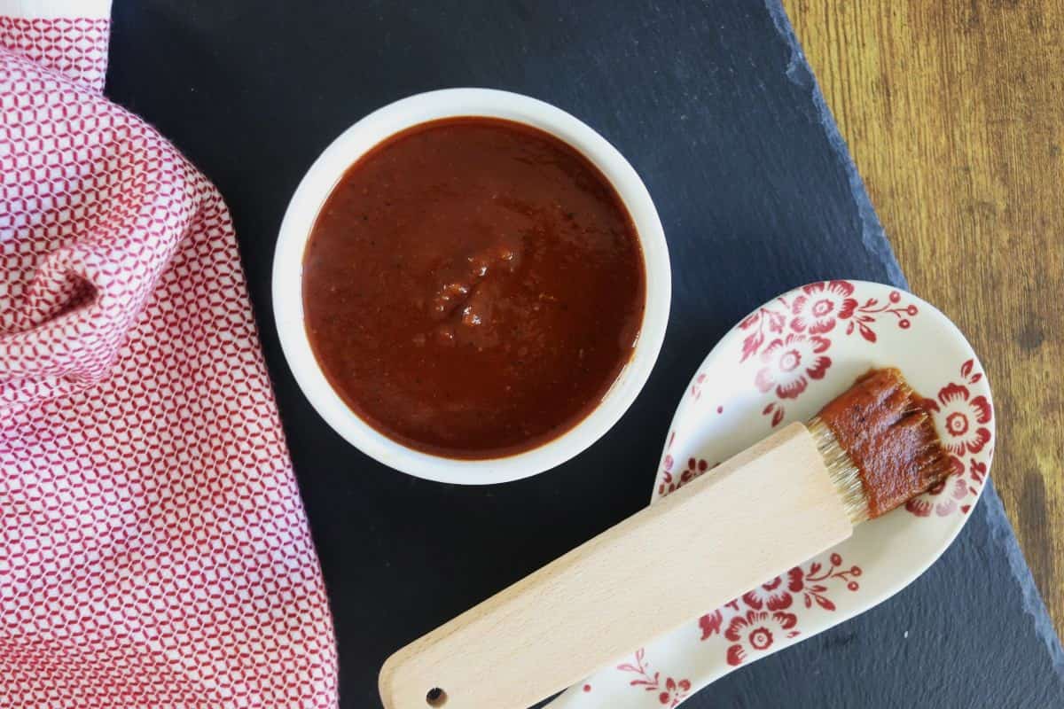 A bowl of dark red barbecue sauce sits on a black slate surface beside a red patterned spoon holder and brush, with a red and white cloth nearby.