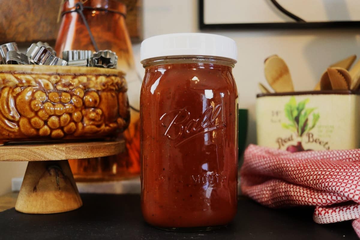 A glass mason jar filled with red tomato sauce sits on a dark surface. Nearby are a brown ceramic bowl with metal cookie cutters, a red-and-white towel, and a tin can with utensils in the background.