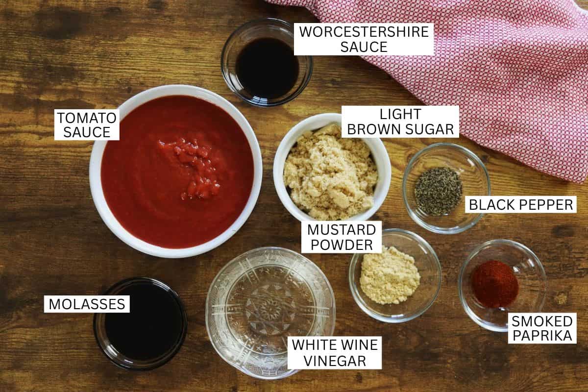 Various BBQ sauce ingredients in bowls on a wooden surface, including tomato sauce, Worcestershire sauce, molasses, light brown sugar, mustard powder, white wine vinegar, black pepper, and smoked paprika.