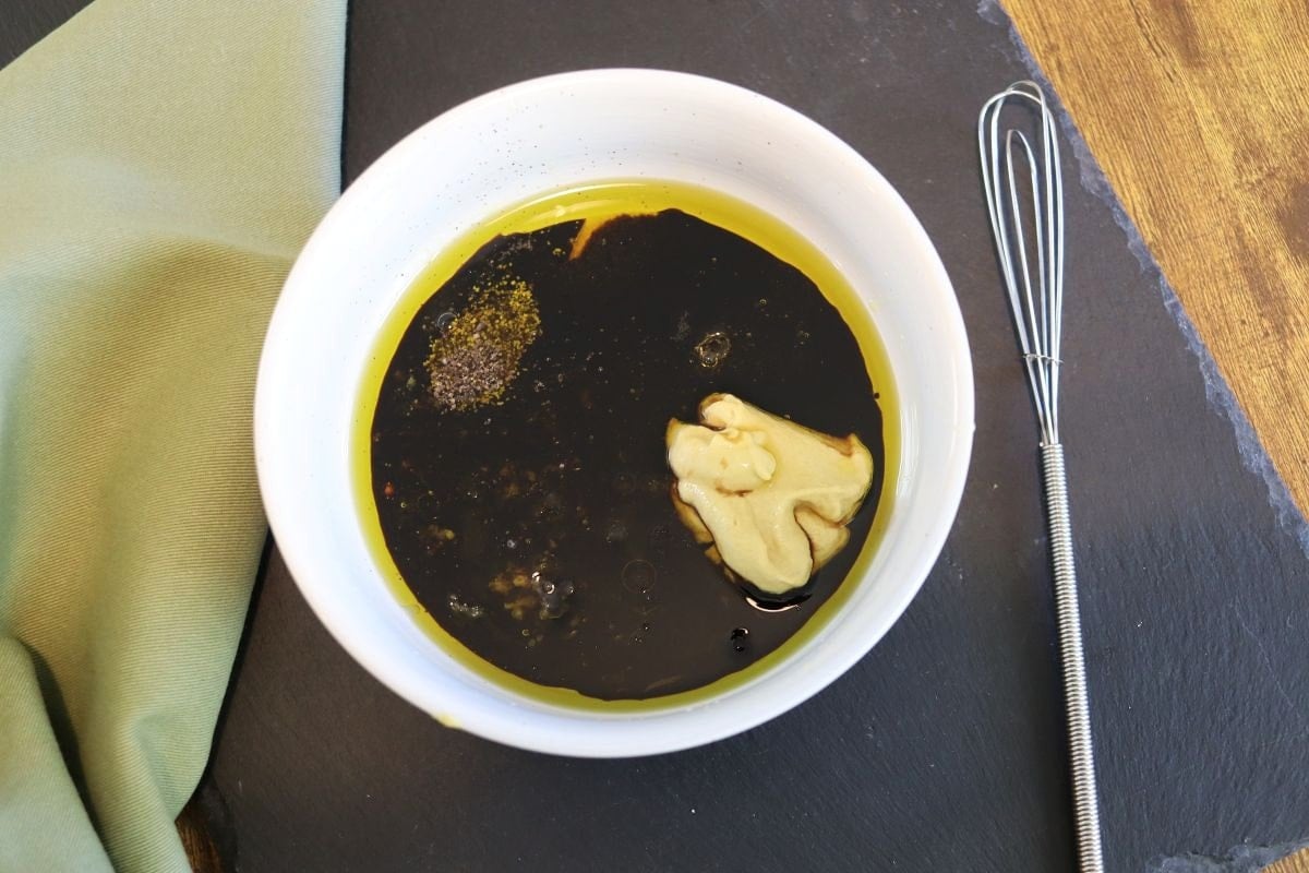 White bowl with dark liquid, yellow oil, dijon mustard, and seasonings in it sitting on a black slate board with a whisk on one side and a green cloth on the other.