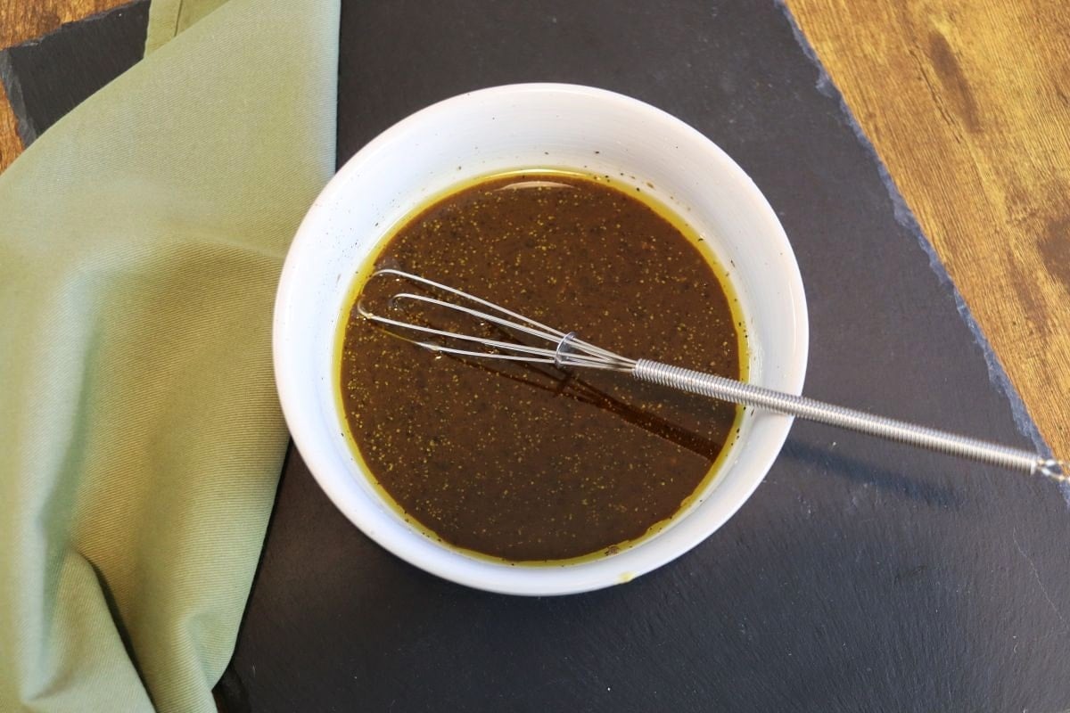 White bowl with dark liquid that has visible spices in it with a whisk in it sitting on a slate board next to a green cloth.