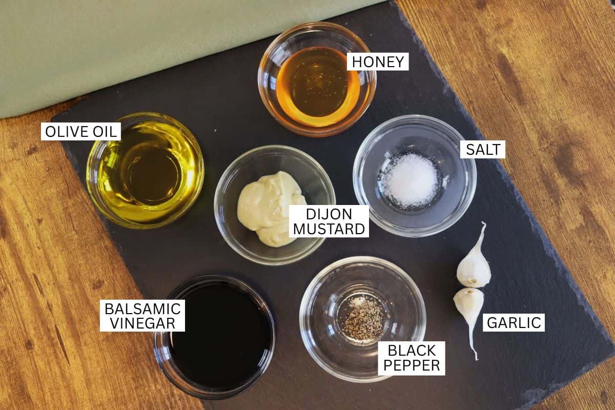 Six labeled small bowls with ingredients for vinaigrette—olive oil, honey, salt, Dijon mustard, balsamic vinegar, and black pepper—are arranged on a black slate with two garlic cloves beside them.