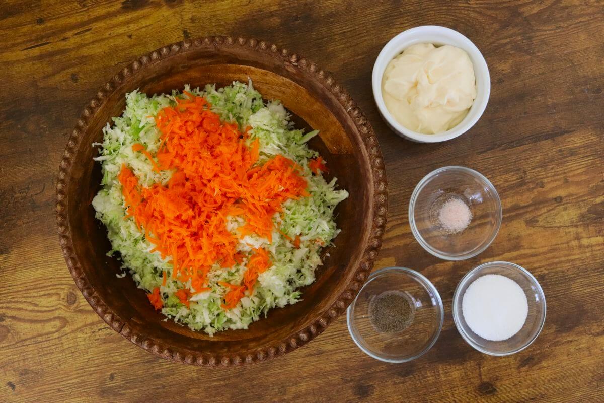 Grated cabbage and carrots in a wooden bowl with small bowls of mayonnaise, sugar, salt, and pepper nearby.