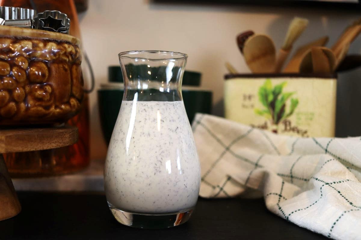 White dressing with herbs visible in it in a glass carafe on a table with a green checkered towel and home decor visible in the background.