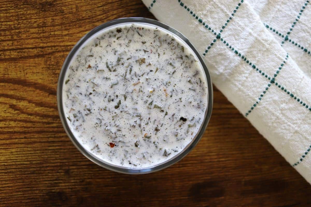 A bowl of creamy dill dressing with visible herbs and spices sits on a wooden surface next to a white towel with green grid lines.