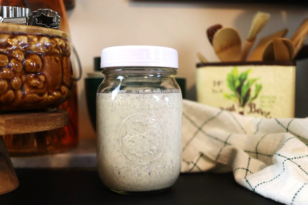 Mason jar of white liquid sitting on a countertop next to a green and white cloth with kitchen utensils in the background.