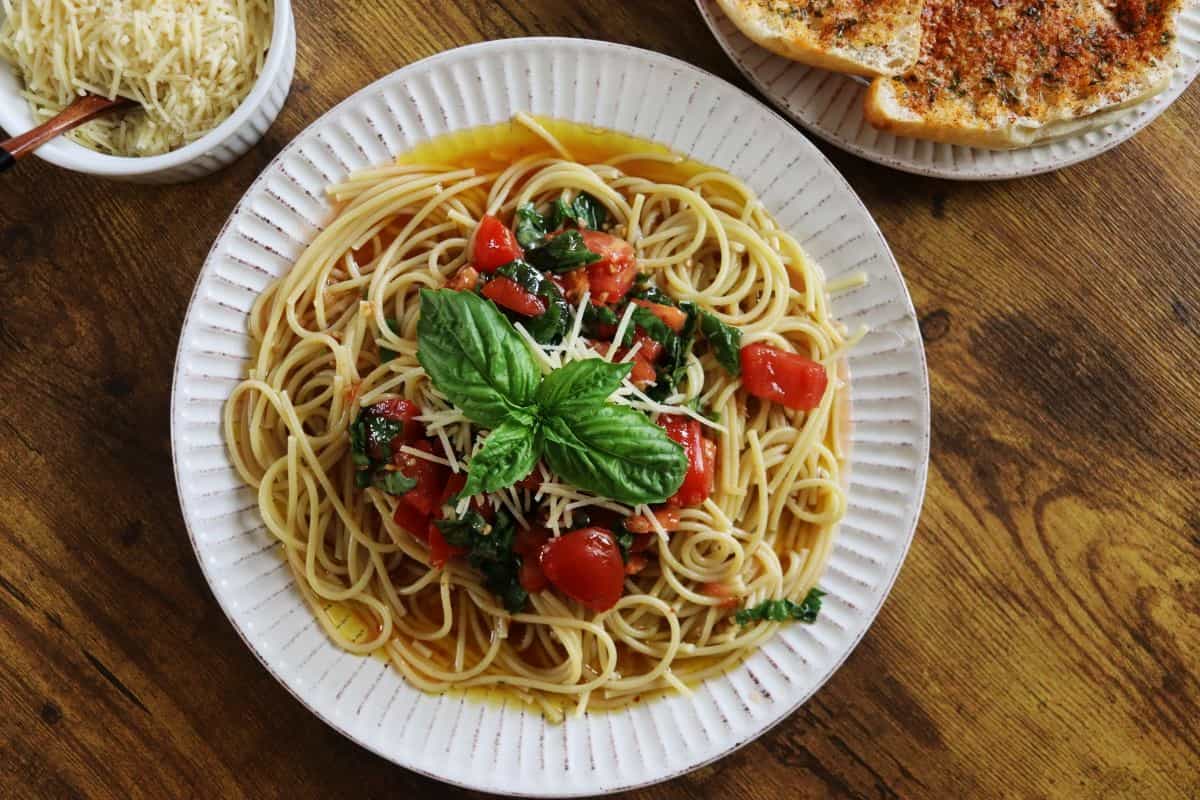 A plate of spaghetti topped with diced tomatoes, fresh basil, and grated cheese sits on a wooden table, with a bowl of shredded cheese and a plate of garlic bread nearby.