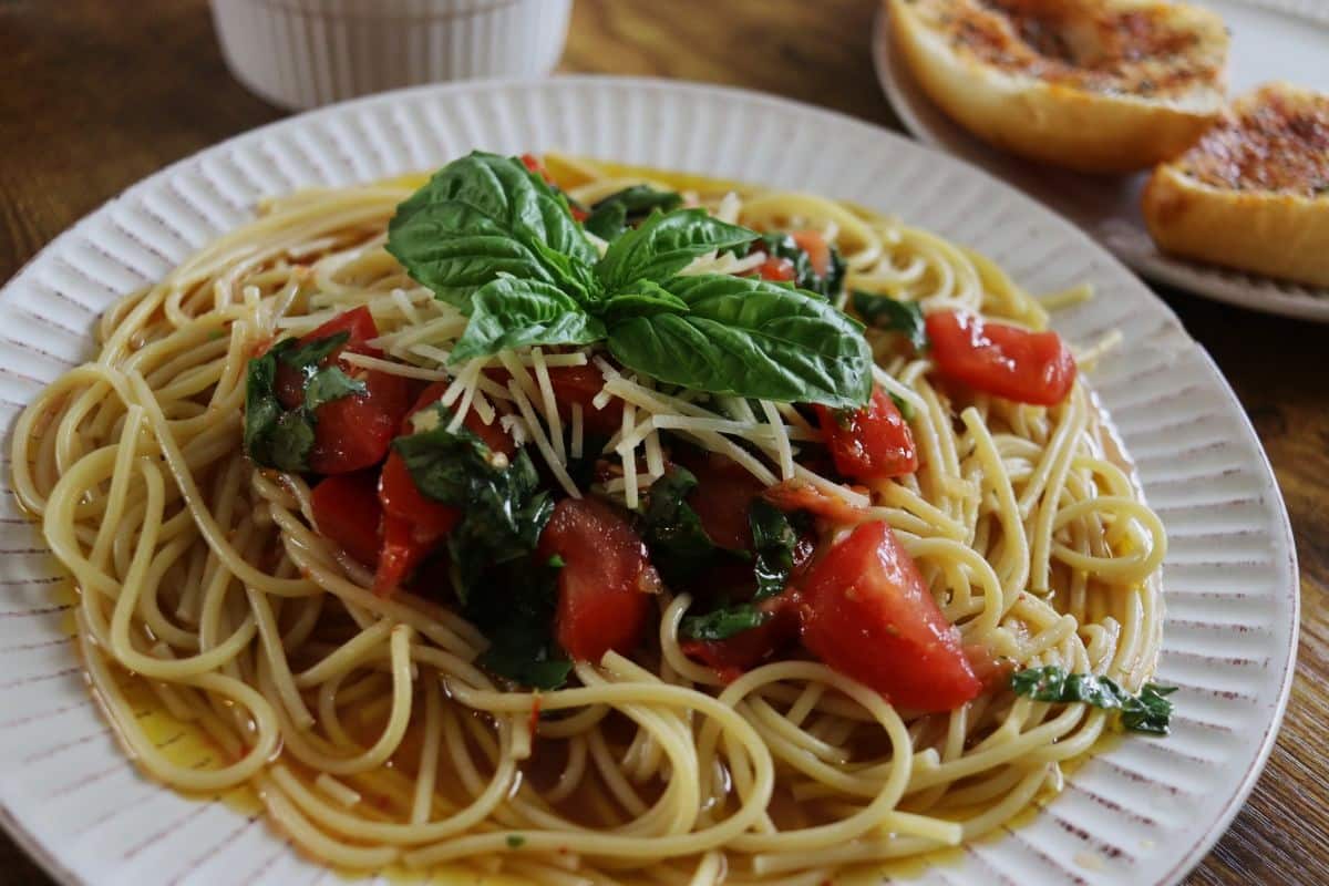 A plate of spaghetti topped with diced tomatoes, fresh basil leaves, and grated cheese, with slices of toasted garlic bread in the background.