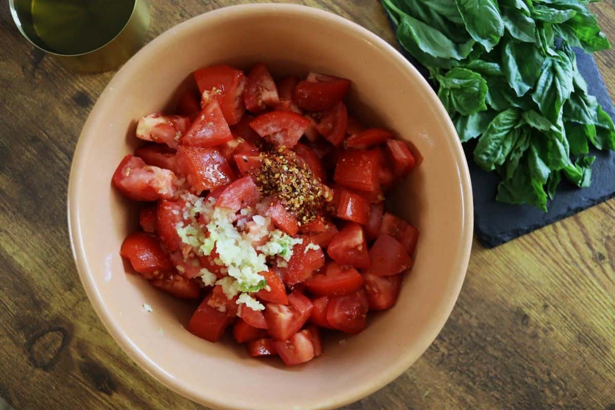 Bowl of chopped tomatoes, pressed garlic, and red pepper flakes sitting on a wooden countertop next to a pile of fresh basil.