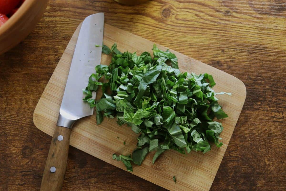 Chopped fresh basil on a small cutting board with a knife on it sitting on a wooden table.