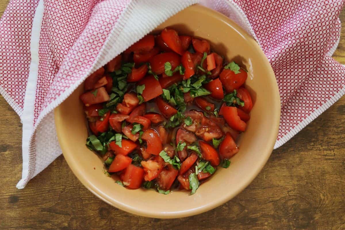Bowl of chopped tomatoes, chopped basil, and oil on a wooden table with a red and white towel by it.