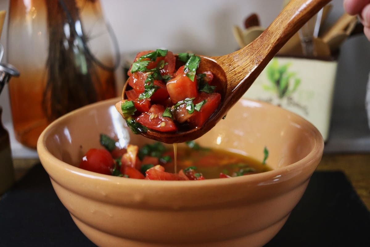 Wooden ladle taking a scoop of tomatoes, basil, and oil out of a bowl with kitchen utensils visible in the background.