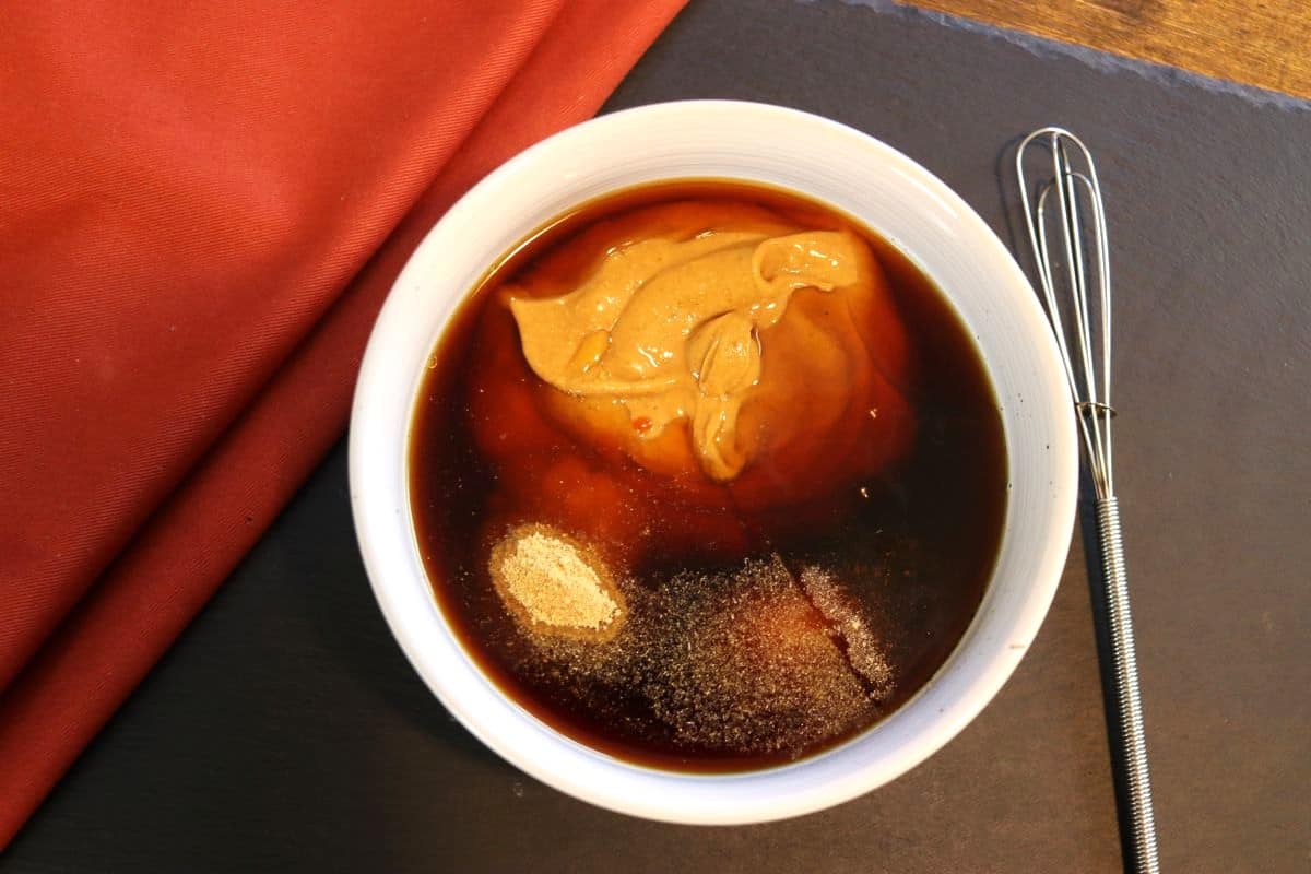 Peanut butter, dark liquid, and spices in a bowl on a slate board with an orange cloth and a whisk next to it.