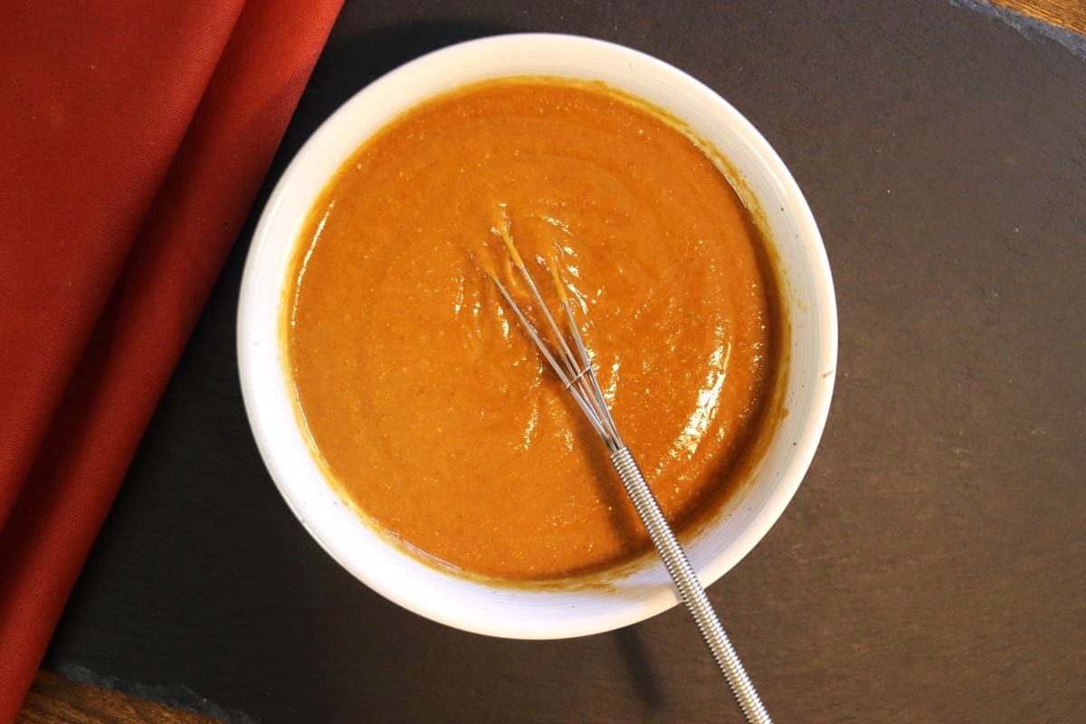 Light brown sauce in a small white dish with a whisk in it sitting on a dark surface.