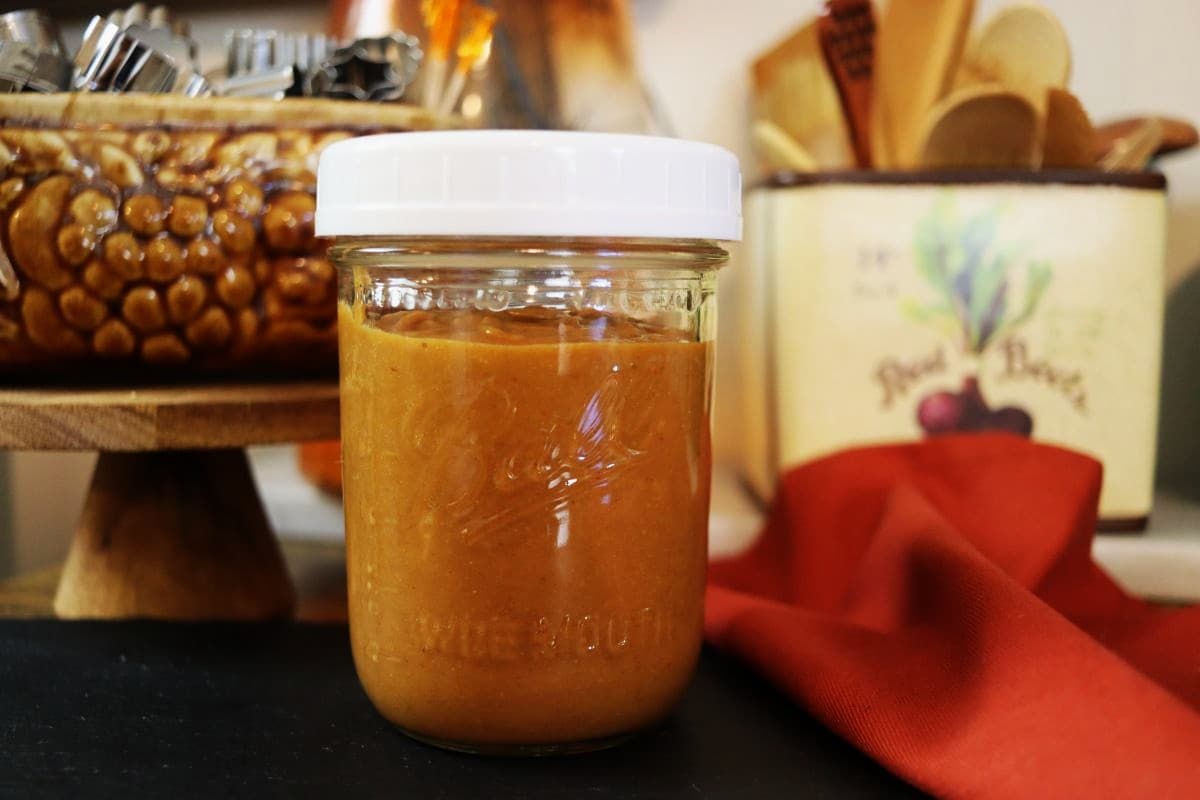 A glass jar with a white lid filled with thick, light brown peanut butter dressing sits on a counter, with a red cloth and kitchen utensils in the background.