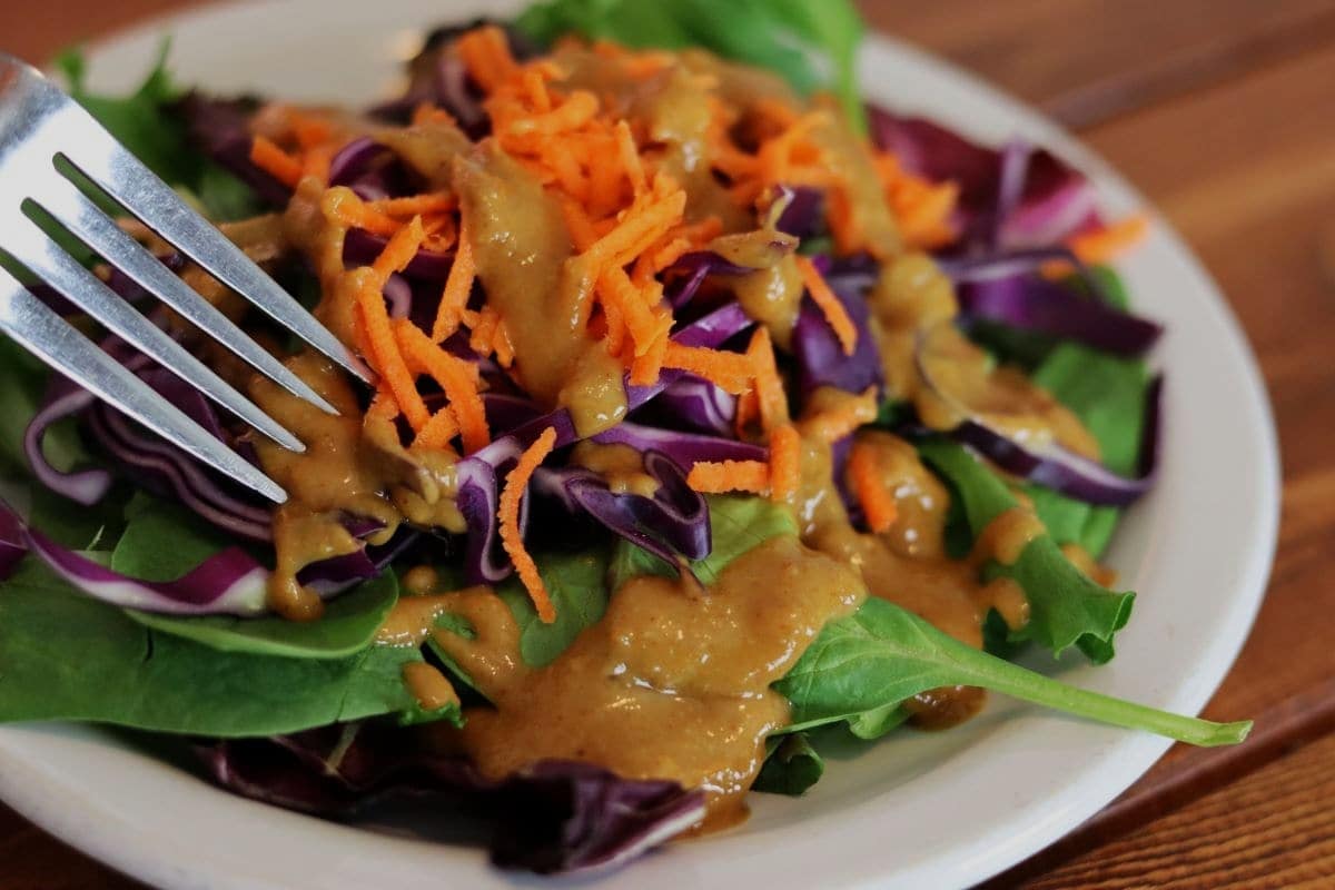 A close-up of a salad with leafy greens, shredded purple cabbage, and grated carrots, topped with a creamy brown dressing, served on a white plate with a fork.