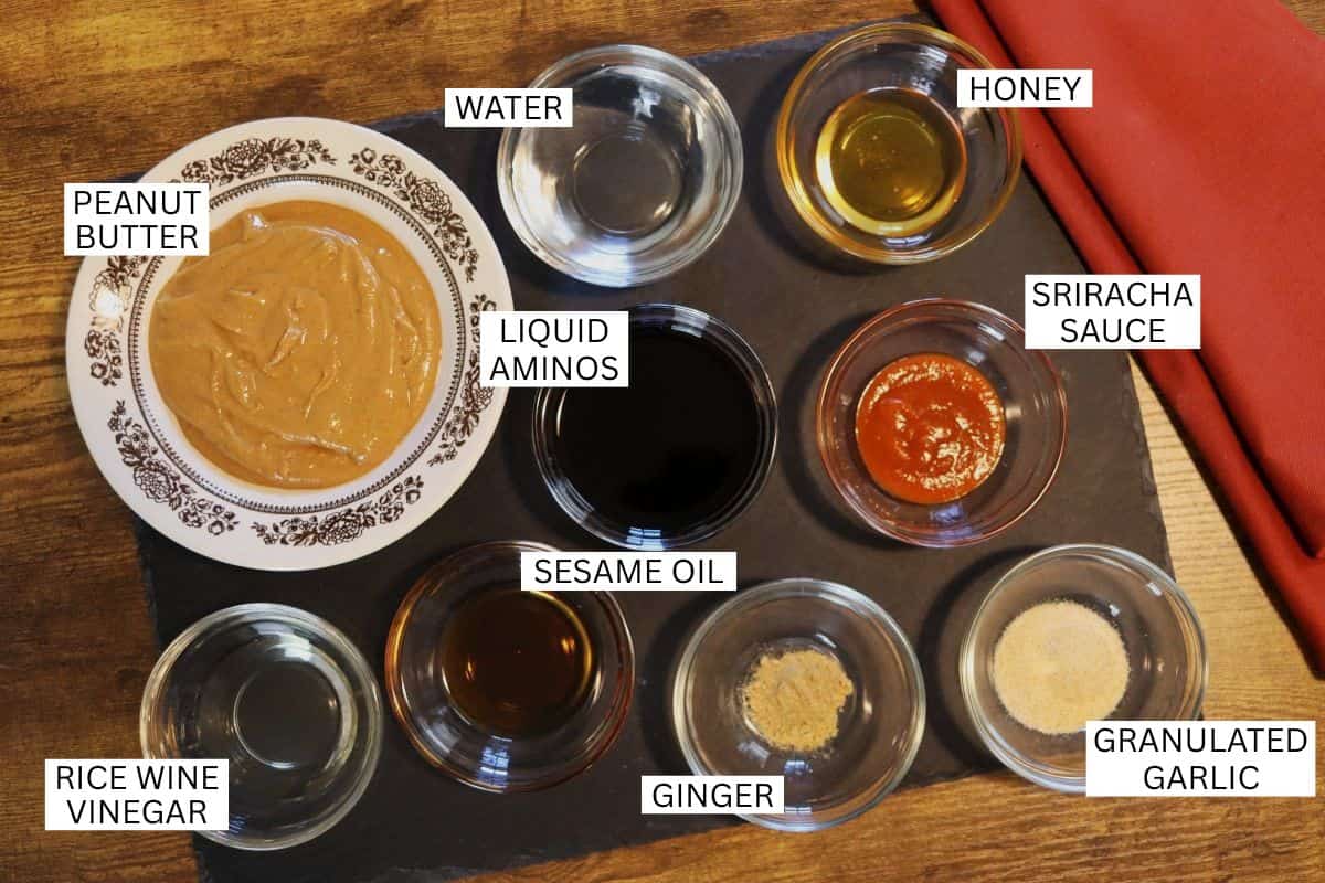 A top view of bowls containing peanut butter, water, honey, sriracha sauce, liquid aminos, sesame oil, rice wine vinegar, ginger, and granulated garlic, all labeled on a dark surface.