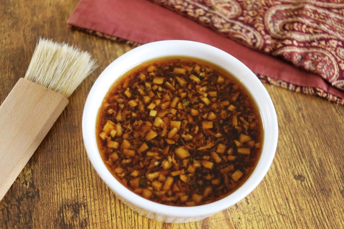 A white bowl filled with a chunky, brown marinade sits on a wooden surface next to a basting brush and a folded, patterned cloth napkin.