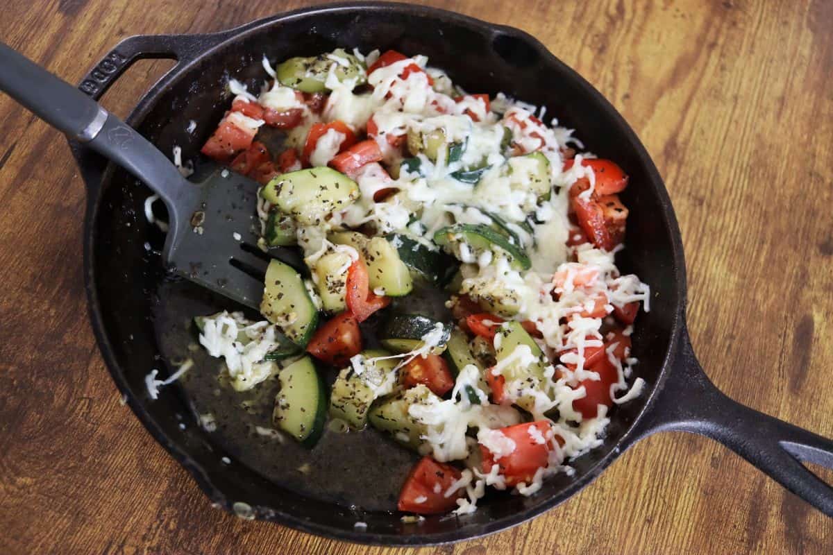 A cast iron skillet on a wooden surface contains sautéed zucchini, tomatoes, melted cheese, and herbs, with a spatula resting inside.
