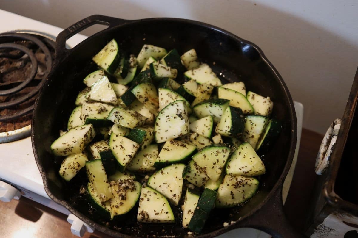 Cut up zucchini covered with seasoning in a cast iron skillet on a stovetop burner.