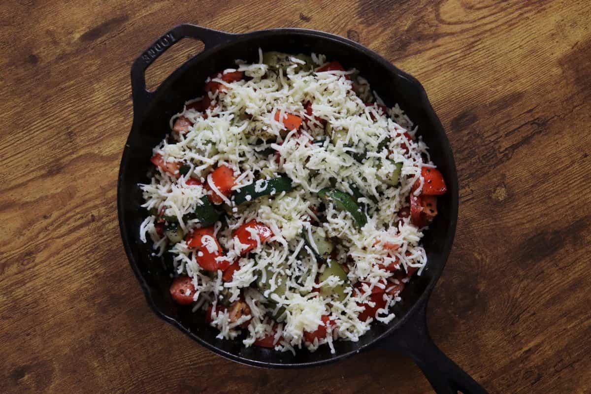 Zucchini and tomatoes topped with shredded mozzarella cheese in a cast iron skillet on a wooden countertop.