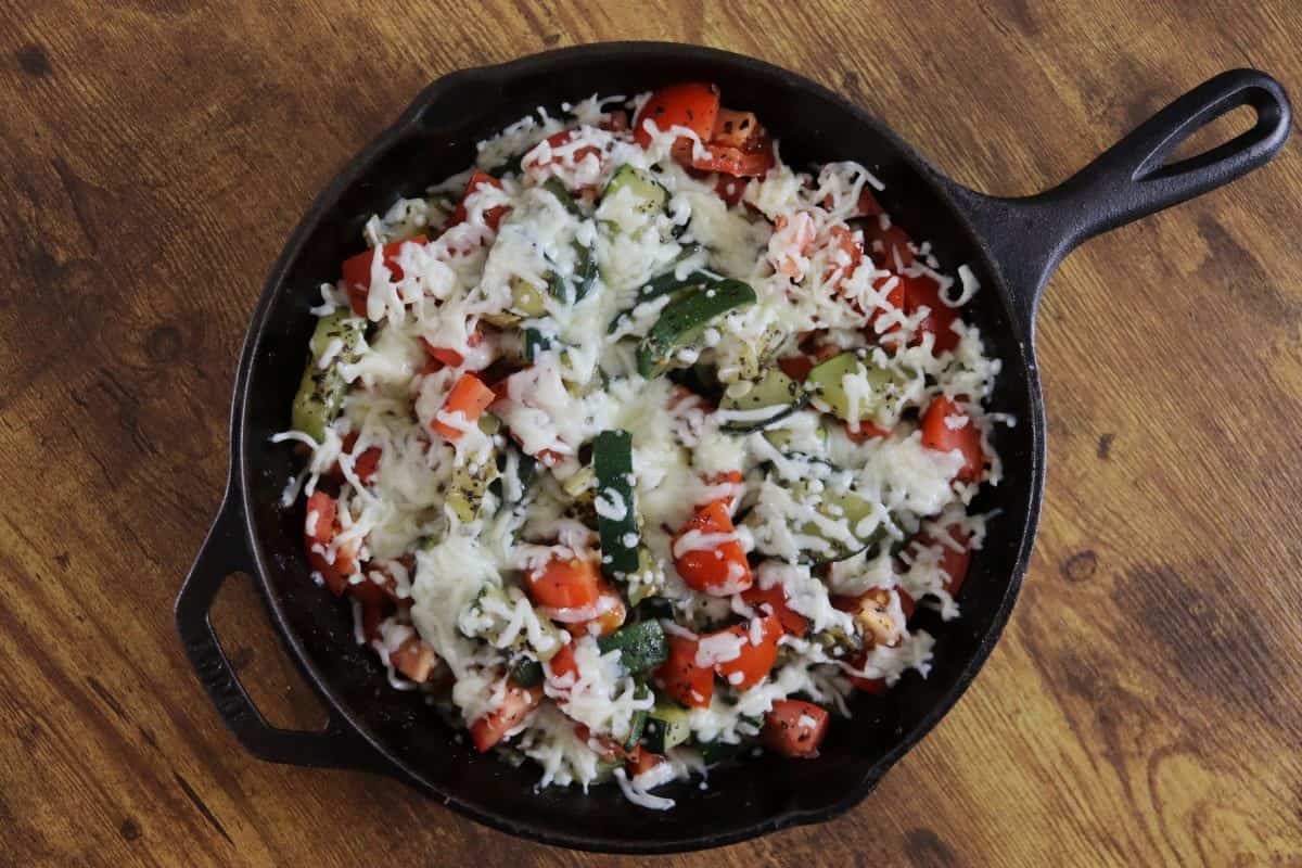 Zucchini and tomatoes topped with melted mozzarella cheese in a cast iron skillet on a wooden countertop.