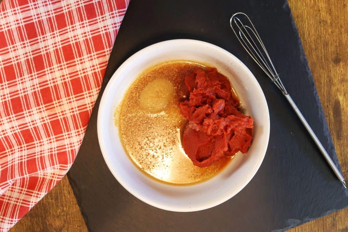Tomato paste, egg, spices, and yellow liquid in a white bowl on a black slate board with a whisk and a red and white checkered cloth next to it.