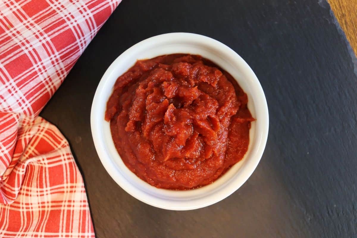 A white bowl filled with thick red ketchup sits on a dark slate surface next to a red and white plaid cloth.
