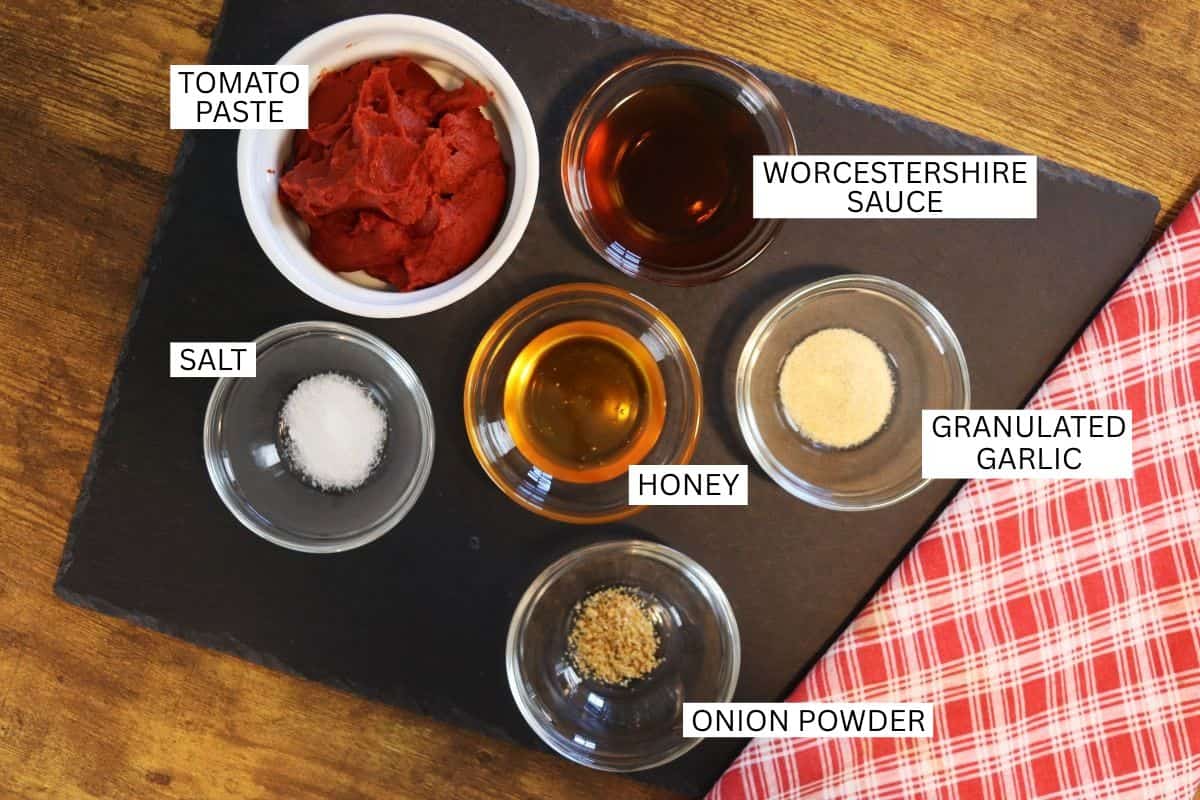 Six small bowls containing tomato paste, Worcestershire sauce, salt, honey, granulated garlic, and onion powder are arranged on a black surface next to a red and white checkered cloth. Each bowl is labeled.