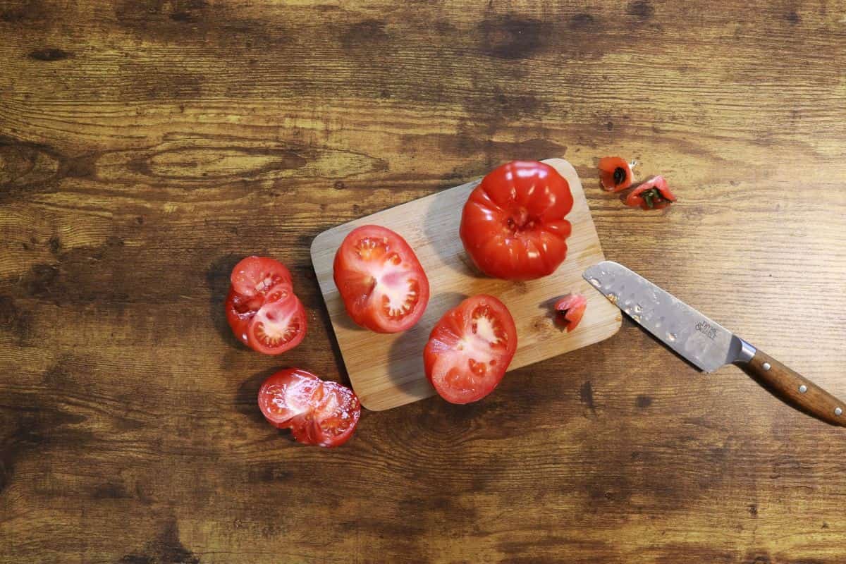 small cutting board on a table with a tomato and tomato halves on it and a knife sitting next to it with tomato cores scattered by the side.