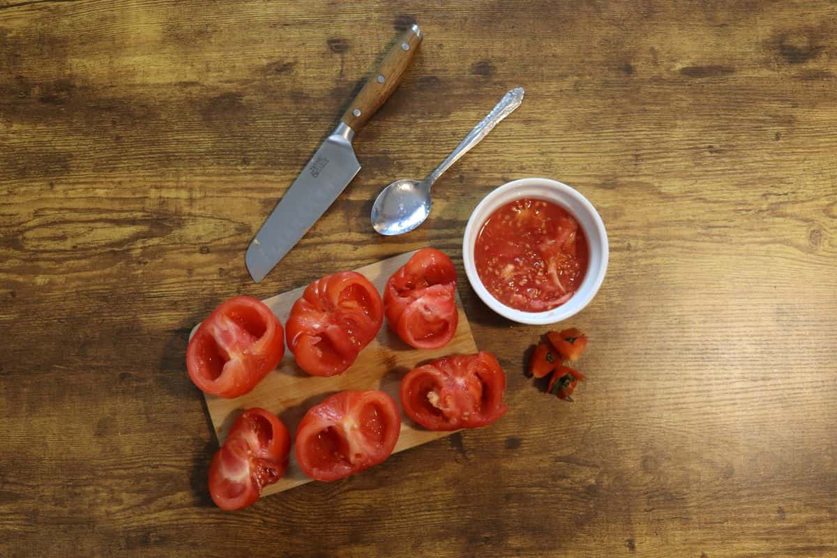 small cutting board on a table with tomato halves on it and a knife, spoon, tomato cores, and a small dish of pulp sitting next to it.