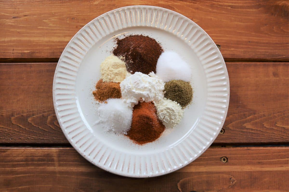 Assorted ground spices and cornstarch arranged in small piles on a white plate set on a wooden table.
