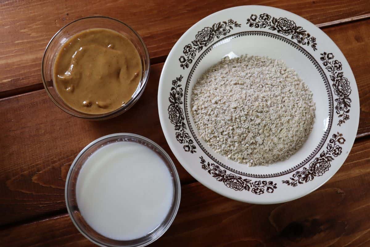 Three bowls on a wooden surface: one with creamy peanut butter, one with oat flour in a patterned bowl, and one with milk. Each bowl is labeled with its ingredient.