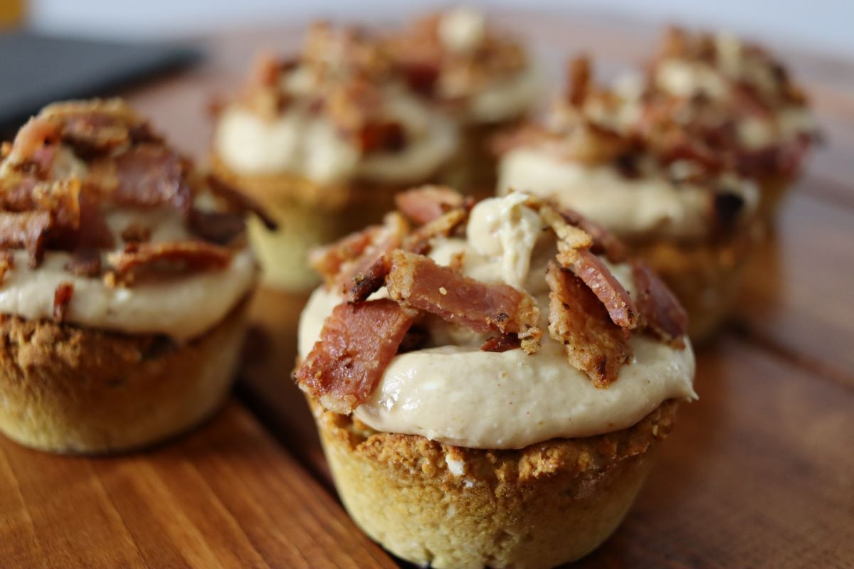 Dog treat that looks like a muffin topped with white icing and chopped bacon on a wooden surface with others visible in the background.