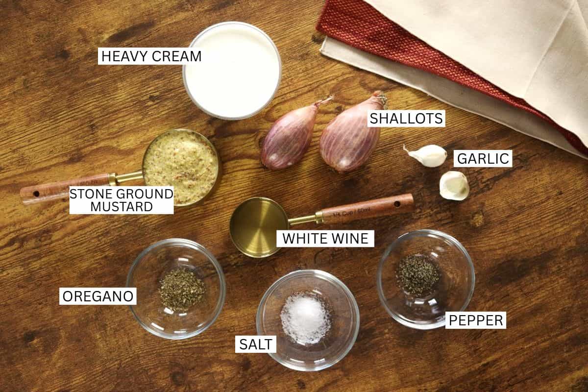 Ingredients for a sauce are arranged on a wooden surface: heavy cream, stone ground mustard, shallots, garlic, white wine, oregano, salt, and pepper, each labeled with white text. A red napkin is in the corner.