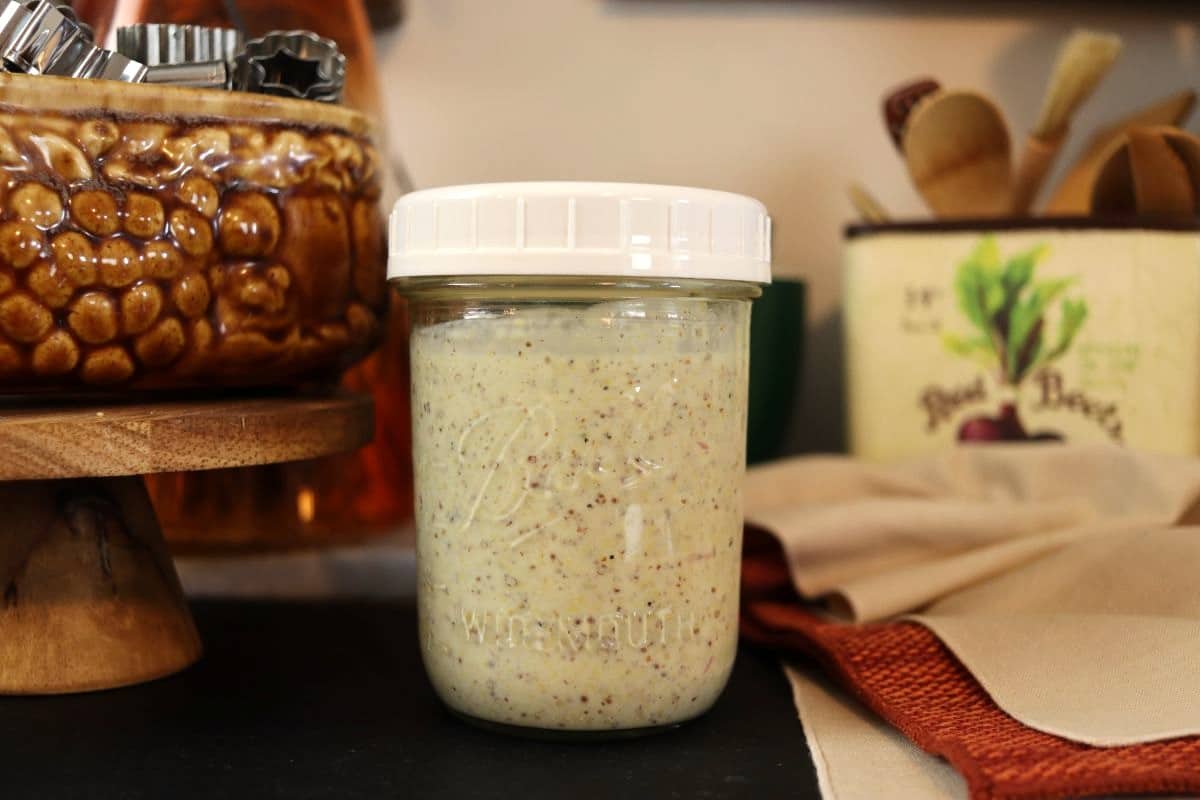 A glass jar with a white lid filled with creamy homemade stone ground mustard sauce sits on a kitchen counter, surrounded by a brown decorative container and utensils in the background.