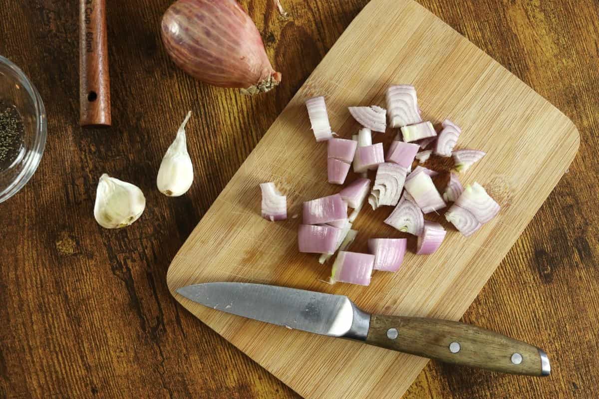 Chopped shallots on a small cutting board with a knife sitting on it and garlic cloves and a whole shallot on the side.