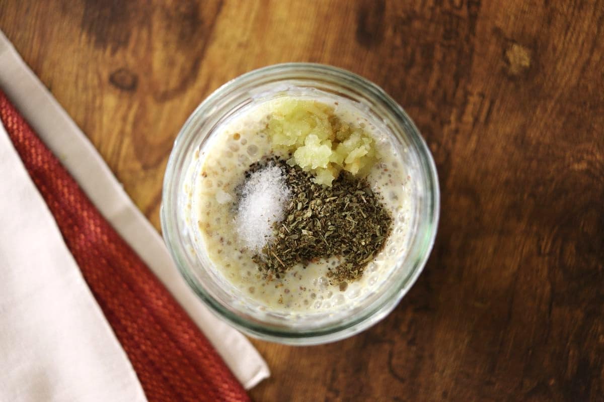 Yellow liquid in a mason jar with spices sitting on top of it on a counter next to tan and orange cloths.