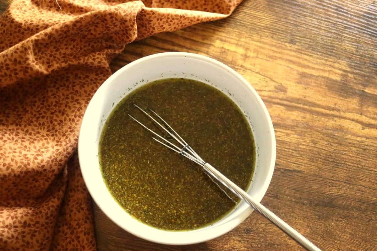 Brownish yellow viniagrette with herbs visible in it in a bowl with a whisk on a table with a brown patterned cloth next to it.
