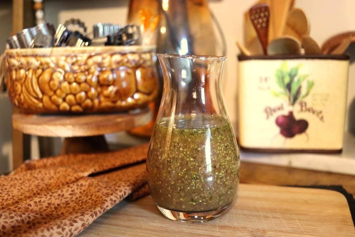 A glass carafe filled with vinaigrette sits on a wooden surface, with a patterned brown cloth nearby. In the background are a ceramic crock with kitchen utensils and a container decorated with beets.