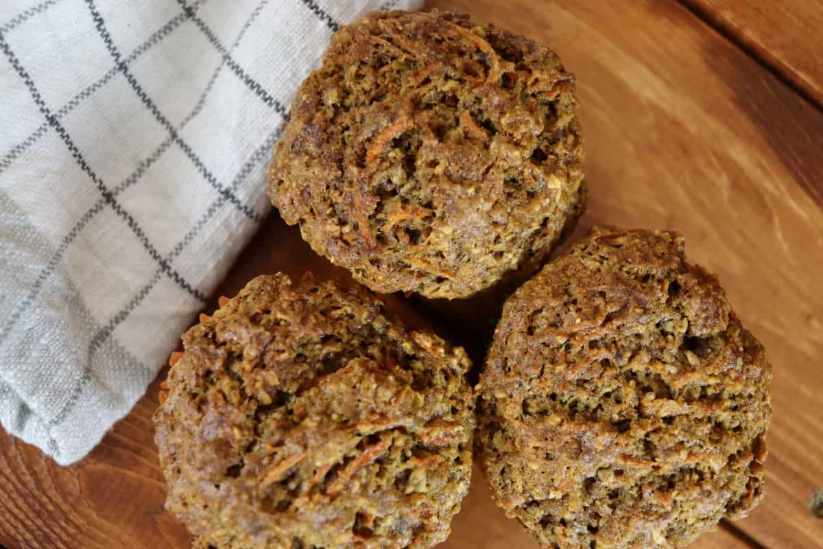 Three golden brown carrot muffins sitting on a wooden surface with a green checkered cloth next to them.
