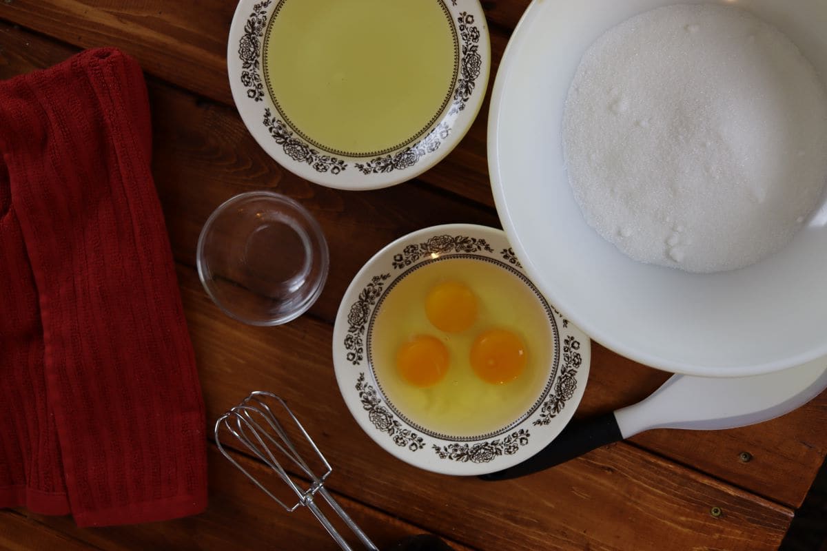Bowls with oil, sugar, and eggs sitting on a counter with a set of beaters next to them.