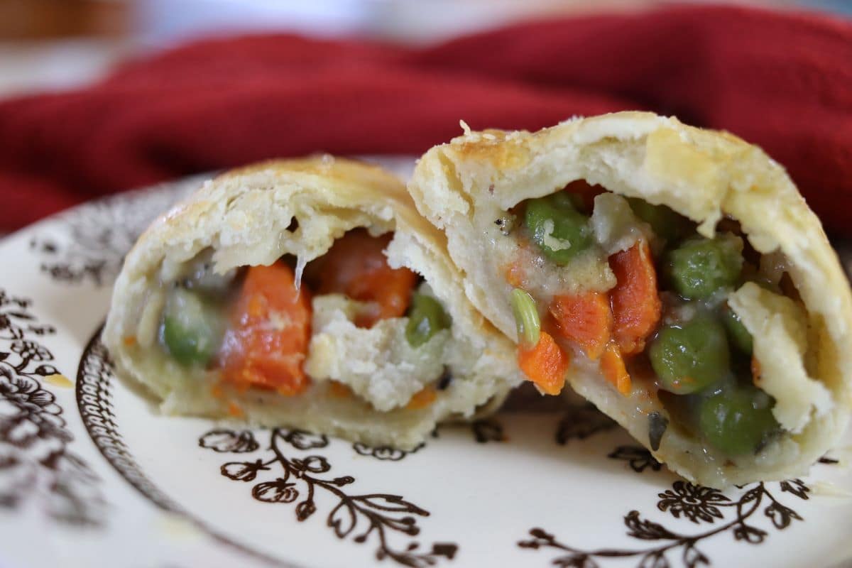 Flaky pastry cut in half on a decorative plate, revealing a filling of mixed vegetables including peas and carrots. A red cloth is blurred in the background.