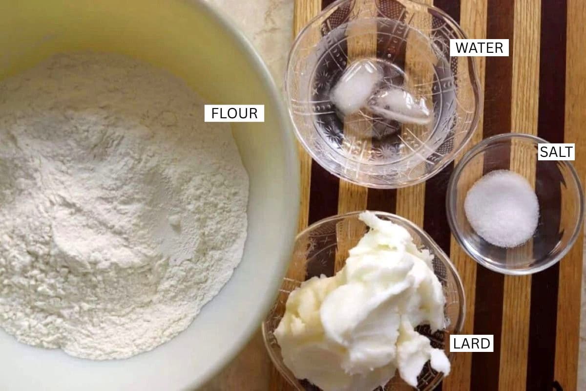 A bowl of flour, and small bowls of lard, salt, and water with ice, are arranged on a wooden striped surface. Each ingredient is labeled.
