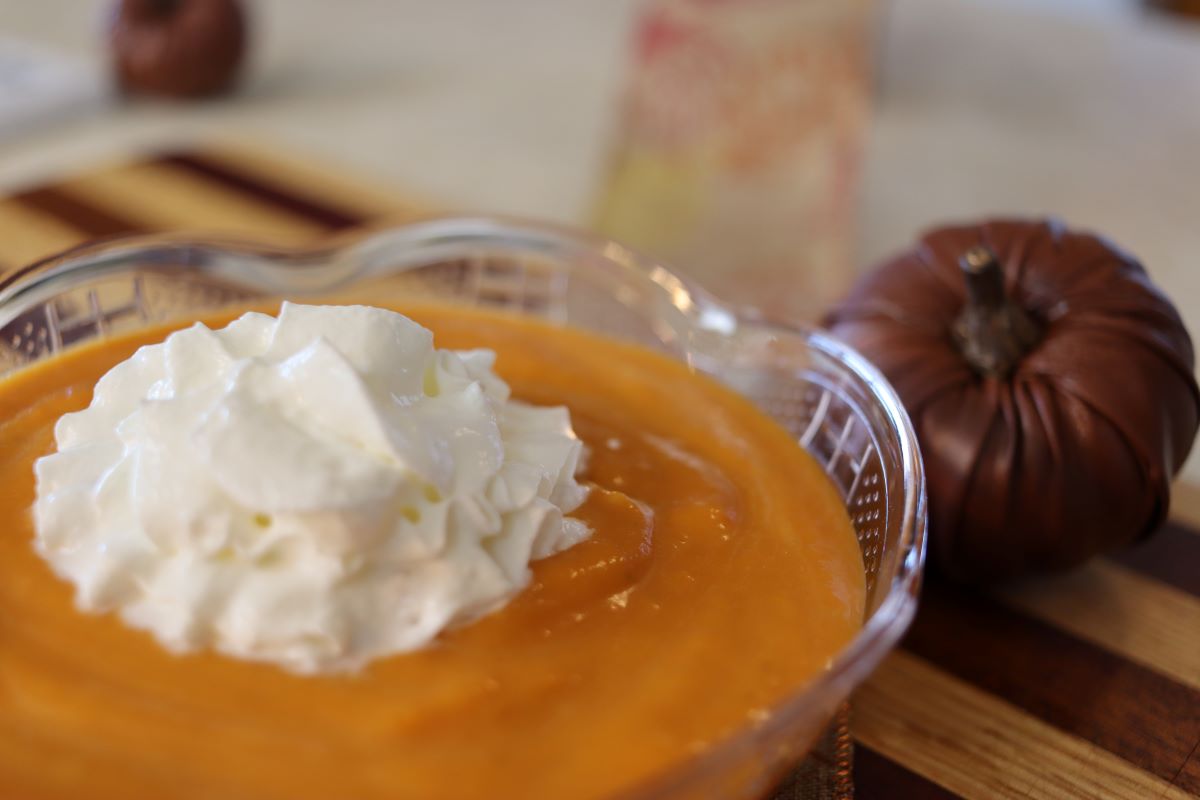 A clear glass bowl filled with pumpkin latte for dogs, topped with a dollop of whipped cream. A decorative brown pumpkin and a blurred background are visible behind the bowl.